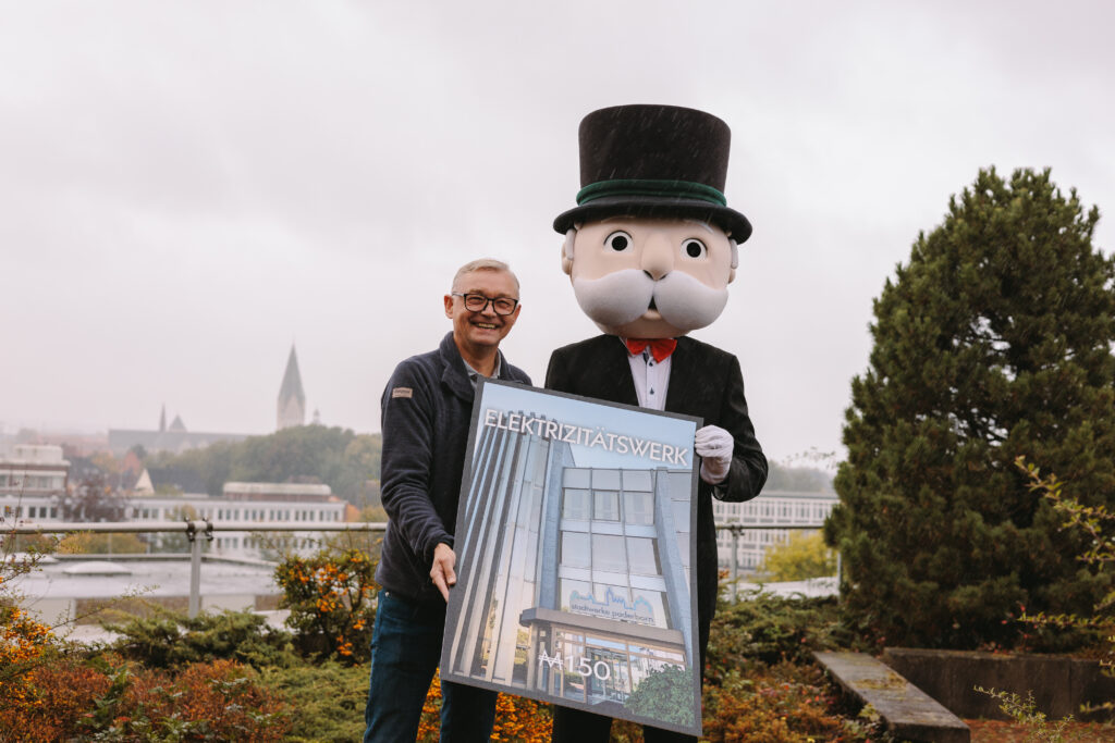 Geschäftsführer Christian Müller reicht Monopoly Maskottchen präsentieren ein Poster mit dem Schriftzug Elektrizitätswerk