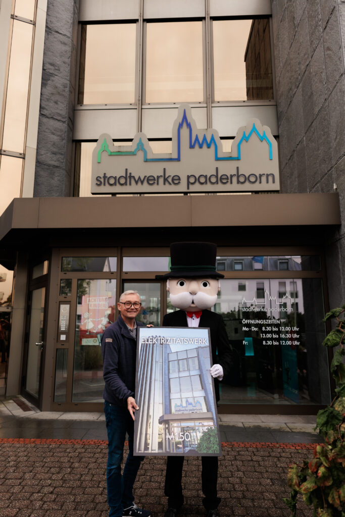 Geschäftsführer Christian Müller reicht Monopoly Maskottchen präsentieren ein Poster mit dem Schriftzug Elektrizitätswerk vor dem Eingang der Stadtwerke Paderborn