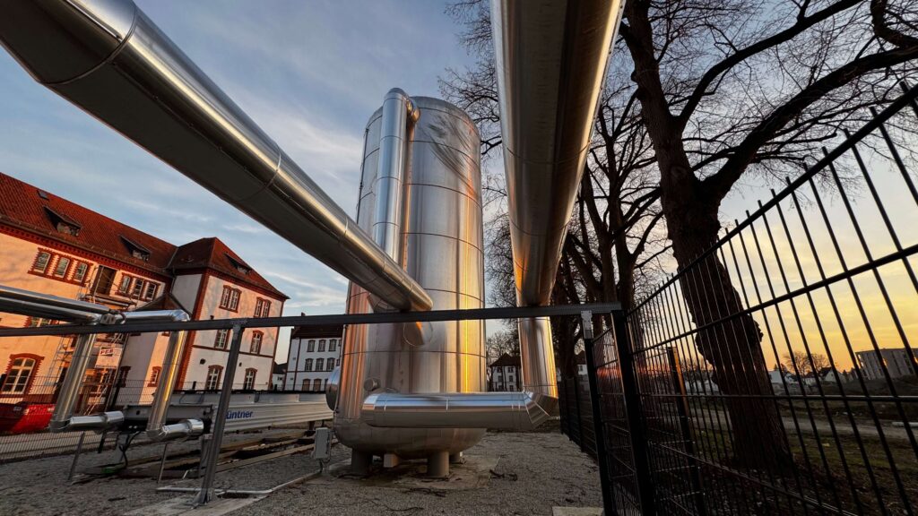 Nahaufnahme einer modernen, silbern isolierten Fernwärme- oder Industrieanlage mit großen Rohrleitungen, die zwischen Gebäuden und Bäumen im warmen Licht des Sonnenuntergangs verlaufen.