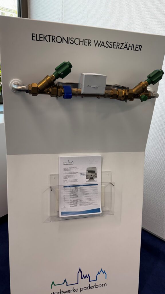 Ausstellungsmodul mit einem eingebauten elektronischen Wasserzähler und Absperrventilen, beschriftet mit „Elektronischer Wasserzähler“, darunter eine Bedienungsanleitung der Stadtwerke Paderborn.